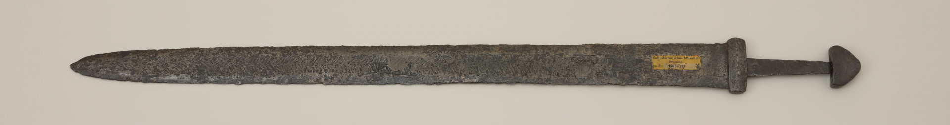 broń; miecz dwusieczny - Ujęcie tyłu miecza. Całkowicie zachowany. Dwusieczna głownia jest szeroka i masywna, o mniej więcej jednakowej szerokości na całej długości, przy zastawie ma przekrój sześcioboczny. Sztych jest zaokrąglony. Na obu płazach głowni widoczny wzór w postaci „jodełki” świadczący o wieloelementowej jej konstrukcji. Rdzeń został wykonany z dwóch skręconych prętów, nisko- i wysokowęglowego, tworzących jądro, z dodanymi po obu stronach nakładkami ostrzy. Trzpień miecza jest wąski i lekko zwęża się ku głowicy. Jelec jest krótki i wysoki, o kształcie widzianym od góry zbliżonym do prostokąta o lekko wypukłych krótszych bokach. Głowica jest jednolita, zbliżona do trójkąta. Jelec i głowica są lite, wykute każde z pojedynczego kawałka metalu.