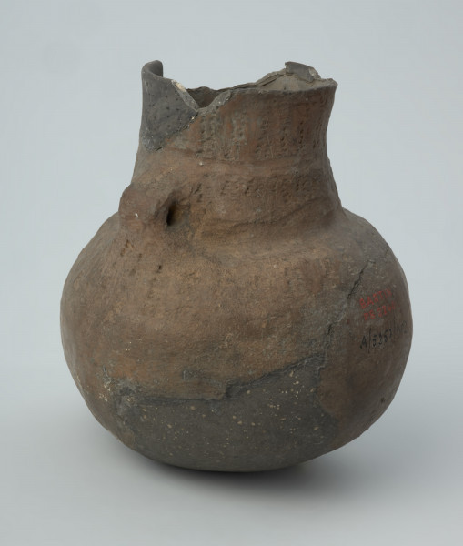 Naczynie gliniane - Ujęcie z prawego boku; Amfora o baniastym brzuścu i cylindrycznej szyjce z parą taśmowatych uch umieszczonych pomiędzy nasadą szyjki i dolną jej partią, zdobiona na szyjce i w górnej części brzuśca trzema dookolnymi pasmami pionowych i ukośnych słupków wykonanych w technice ściegu bruzdowego, rozdzielonych w połowie wysokości szyjki dookolną bruzdą oraz szeroką bruzdą ze śladami wygniatania palcami na przejściu szyjki w brzusiec. Naczynie zrekonstruowane i uzupełnione gipsem barwionym. Barwa od szarobrunatnej do brązowej.