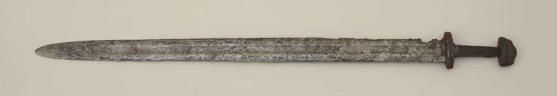 miecz dwusieczny - Ujęcie przodu miecza z góry. Egzemplarz całkowicie zachowany, z korozyjnymi ubytkami. Głownia posiada szerokie zbrocze, widoczne nieomal na całej jej długości. Została wykonana w technice prętowej, metodą dziwerowania. Ślady zasosowania tej techniki widoczne są po obu stronach głowni, z jednej w postaci tzw. „jodełki”, a z drugiej strony jako tzw. „dziwer gwiaździsty”. Świadczy to o użyciu dwóch lub czterech elementów, z których każdy wykonany został z siedmiowarstwowych laminowanych prętów, następnie poddanych skręceniu i finalnie przepołowionych. Następnie dodano nakładki ostrzowe wykonano z warstwowanych taśm o różnym stopniu nawęglania. Rękojeść miecza ma krótki, masywny jelec, który w przekroju bocznym przypomina ośmiobok, oraz dwuczęściową głowicę. Trzpień do rękojeści ma kształt zbliżony do prostokąta i nieznacznie zwęża się ku głowicy. Odkryto na nim ślady organicznych okładzin. Elementy oprawy pierwotnie były ornamentowane w postaci cienkich pionowych pasków wykonanych ze stopu miedzi i cynku.