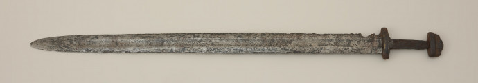 miecz dwusieczny - Ujęcie przodu miecza z góry. Egzemplarz całkowicie zachowany, z korozyjnymi ubytkami. Głownia posiada szerokie zbrocze, widoczne nieomal na całej jej długości. Została wykonana w technice prętowej, metodą dziwerowania. Ślady zasosowania tej techniki widoczne są po obu stronach głowni, z jednej w postaci tzw. „jodełki”, a z drugiej strony jako tzw. „dziwer gwiaździsty”. Świadczy to o użyciu dwóch lub czterech elementów, z których każdy wykonany został z siedmiowarstwowych laminowanych prętów, następnie poddanych skręceniu i finalnie przepołowionych. Następnie dodano nakładki ostrzowe wykonano z warstwowanych taśm o różnym stopniu nawęglania. Rękojeść miecza ma krótki, masywny jelec, który w przekroju bocznym przypomina ośmiobok, oraz dwuczęściową głowicę. Trzpień do rękojeści ma kształt zbliżony do prostokąta i nieznacznie zwęża się ku głowicy. Odkryto na nim ślady organicznych okładzin. Elementy oprawy pierwotnie były ornamentowane w postaci cienkich pionowych pasków wykonanych ze stopu miedzi i cynku.