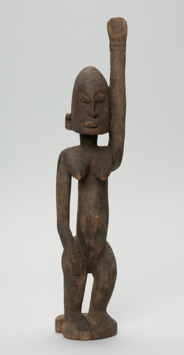 rzeźbiona figura - Ujęcie z przodu. Drewniana, rzeźbiona postać kobiety. Jej lewa ręka podniesiona jest do góry.