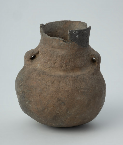 Naczynie gliniane - Ujęcie z przodu; Amfora o baniastym brzuścu i cylindrycznej szyjce z parą taśmowatych uch umieszczonych pomiędzy nasadą szyjki i dolną jej partią, zdobiona na szyjce i w górnej części brzuśca trzema dookolnymi pasmami pionowych i ukośnych słupków wykonanych w technice ściegu bruzdowego, rozdzielonych w połowie wysokości szyjki dookolną bruzdą oraz szeroką bruzdą ze śladami wygniatania palcami na przejściu szyjki w brzusiec. Naczynie zrekonstruowane i uzupełnione gipsem barwionym. Barwa od szarobrunatnej do brązowej.