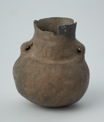 Naczynie gliniane - Ujęcie z przodu; Amfora o baniastym brzuścu i cylindrycznej szyjce z parą taśmowatych uch umieszczonych pomiędzy nasadą szyjki i dolną jej partią, zdobiona na szyjce i w górnej części brzuśca trzema dookolnymi pasmami pionowych i ukośnych słupków wykonanych w technice ściegu bruzdowego, rozdzielonych w połowie wysokości szyjki dookolną bruzdą oraz szeroką bruzdą ze śladami wygniatania palcami na przejściu szyjki w brzusiec. Naczynie zrekonstruowane i uzupełnione gipsem barwionym. Barwa od szarobrunatnej do brązowej.