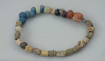 kolia paciorkowa - Ujęcie z góry tylnej części naszyjnika. Kolia 32 paciorków szklanych i 2 paciorków z karneolu. Pierwszy paciorek z karneolu ma kształt kubooktaendryczny, drugi dwustożkowaty, w rzucie z góry ma formę wielokąta. Paciorki szklane są także zróżnicowane: 6 niebieskich jest beczułkowatych lub płasko-kulistych, 3 bezbarwne mają podobne kształty, 2 grafitowe są płasko-kuliste, 1 granatowy, 1 szary z powodu pokrycia patyną, 2 płasko-wypukłe o zielonkawej barwie, 2 płasko-kuliste zdobione nadtapianą nicią tworzącą falisty wzór, 2 dwusegmentowe o nieokreślonym kolorze z powodu spatynowania oraz 13 cylindrycznych w tym 9 kanelurowanych, pokrytych ornamentem powielonej linii falistej. Średnica paciorków waha się między 0,6 a 1,2 cm. Całość jest w różnym stopniu podniszczona, paciorki są w większości matowe, niektóre całkowicie pokryte patyną.