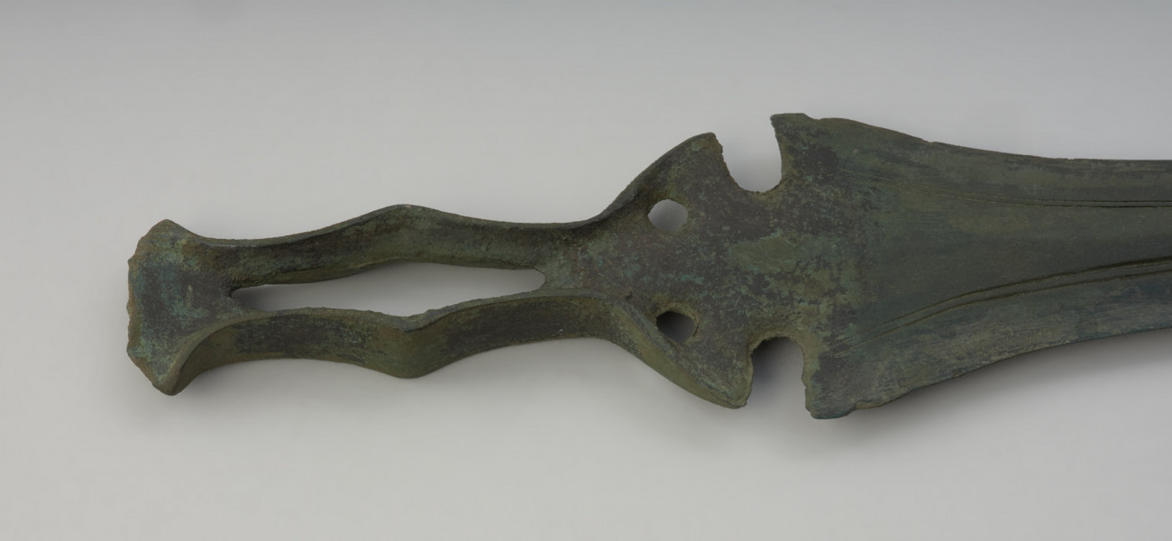 miecz ze sztabą do rękojeści - Ujęcie głowni miecza w poziomie z przodu. Zachowany w całości miecz obosieczny o smukłej głowni z widocznymi podwójnymi struzinami na powierzchni. Sztaba do rękojeści ma oryginalną formę, poszerzoną pośrodku i zwieńczoną wachlarzowatym zakończeniem. Dodatkowo na całej jej długości przebiega charakterystyczna, rombowata szczelina. Pókolista podstawa sztaby zaopatrzona jest w 2 otwory na nity. Obydwie powierzchnie miecza są lekko porowate i pokryte zieloną patyną.