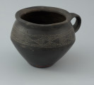 Naczynie gliniane - Ujęcie z przodu. Naczynie gliniane (kubek) posiada gładką, czarną powierzchnię i drobną domieszkę.