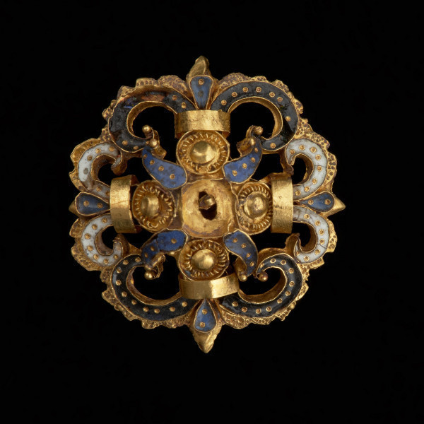 rozeta - Ujęcie z przodu na czarnym tle. Ozdoba w kształcie ażurowej rozety. Wokół centralnie umieszczonego złotego guza rozmieszczone są cztery mniejsze guzy otoczone filigranem, płatki zdobione ciemnoniebieską emalią, a także woluty wypełnione czarną oraz białą emalią, spięte złotymi taśmami. Elementy emaliowane są inkrustowane regularnie rozmieszczonymi złotymi kropkami. Na odwrocie uszko służące do przyszywania rozet do strojów.