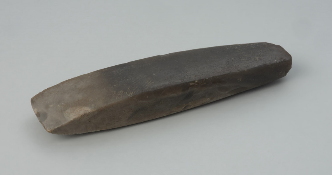 Dłuto krzemienne - Ujęcie na płasko ostrzem w lewą stronę. Dłuto krzemienne, czworościenne, trapezowate, o tępym obuchu. Powierzchnia szlifowana, przy ostrzu i w stronę obucha polerowana do połysku, a na obuchu negatywowa. Krzemień kredowy o barwie od jasno- do ciemnoszarej