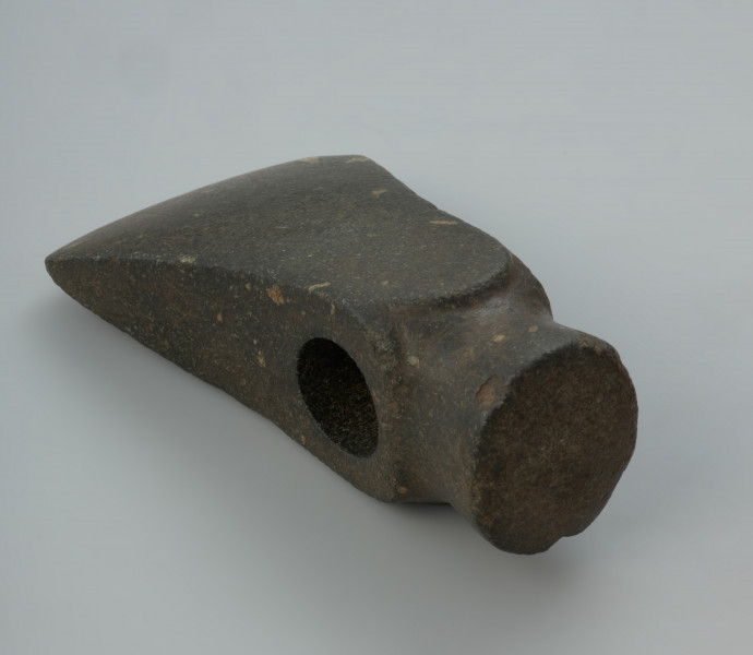 Miniaturowy kamienny toporek. - Ujęcie tyłu. toporka w poziomie. Miniaturowy kamienny toporek z czopowatym obuchem oraz o korpusie lekko rozszerzającym się w kierunku ostrza.