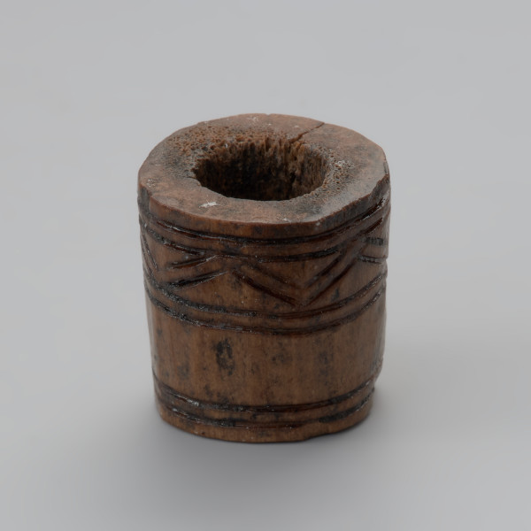 oprawka stożkowata - Ujęcie przodu oprawki w pionie lekko z góry. Oprawka ma stożkowaty kształt o ściętym wierzchołku. Ślady pozostawione na jej powierzchni świadczą o wykorzystaniu do jej produkcji co najmniej trzech narzędzi – piły, noża oraz świdra. Na powierzchni oprawki znajduje się mało skomplikowany wzór zdobniczy o motywie liniowym naniesionym za pomocą ostrego narzędzia. Górną część zajmuje ornament w postaci dwóch dookolnych linii rytych. Natomiast na przeciwległej stronie występuje ozdobnik w formie linii zygzakowatych wpisanych od góry i od dołu w podwójne linie ryte.