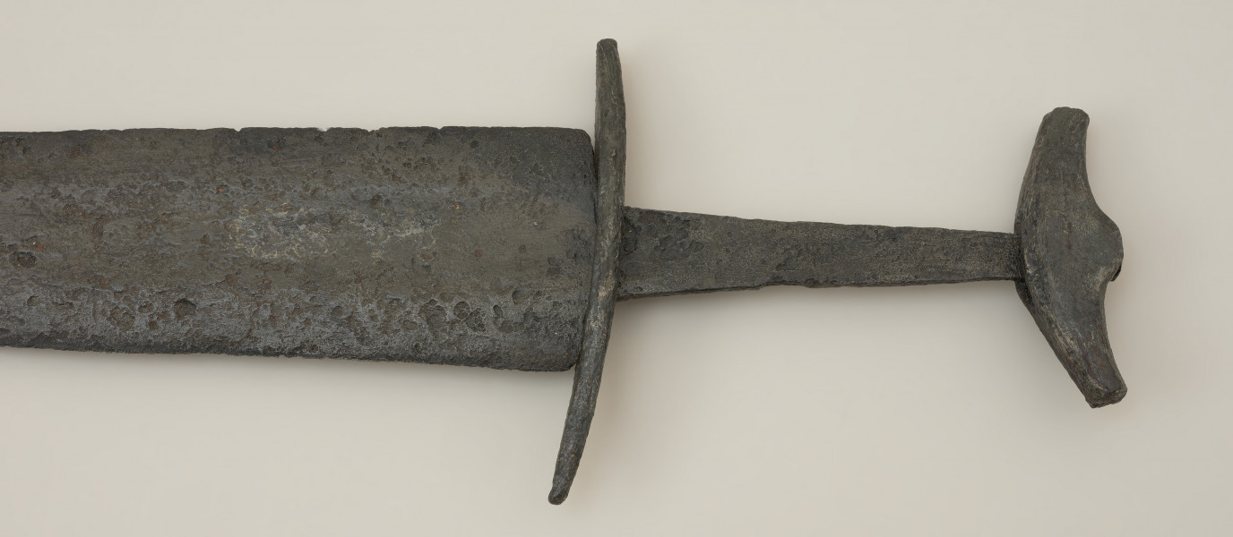 miecz dwusieczny - Ujęcie rękojeści miecza z tyłu. Całkowicie zachowany, z niewielkimi ubytkami korozyjnymi w okolicach sztycha. Jego głownia jest najszersza przy jelcu i równomiernie zwężą się ku ostremu sztychowi. Jest zaopatrzona w szerokie zbrocze. Głownię wykonano w technice prętowej, poprzez zgrzanie stalowych nakładek ostrzowych z jednolitym rdzeniem. Trzpień mocno zwęża się ku głowicy. Jelec, soczewkowaty w rzucie z góry, jest długi i płaski, a jego końce są zakrzywione w kierunku głowni. Jednolita głowica ma kształt siodełkowaty. Jej dolna część wygięta jest ku górze. Na jej powierzchni zaznaczono symboliczny podział na podstawę i nakładkę.