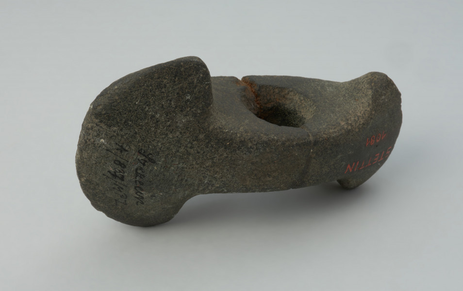 broń obuchowa - Ujęcie na wprost. Topór kamienny obosieczny, tzw. o dwóch ostrzach, labrys lub topór Amazonek, na obu końcach rozszerzony symetrycznie w górę i dół, przy czym koniec ostrzejszy jest słabiej rozchylony, niż druga strona. Otwór do osadzenia trzonka znajduje się około połowy długości korpusu. Przełamany na wysokości otworu i sklejony. Powierzchnia gładzona, nieco zniszczona.