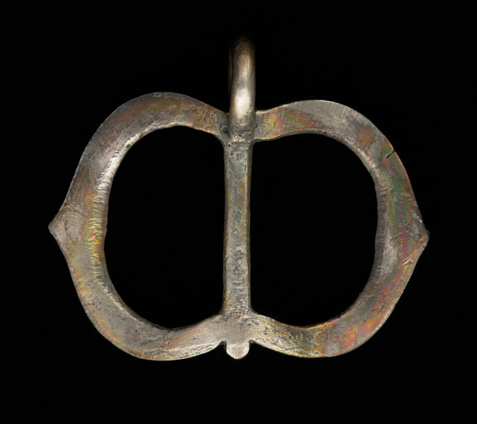 klamra od pendentu - Ujęcie z tyłu. Rama klamry zbudowana z dwóch przedzielonych gładkim trzpieniem części, które w zewnętrznym obrysie mają kształt łuku w ośli grzbiet, a od wewnątrz łuku koszowego. Przednie i boczne ścianki ramy zdobione są rytym, niellowanym ornamentem. U dołu gładkie, okrągłe uszko, mocowane poprzecznie.