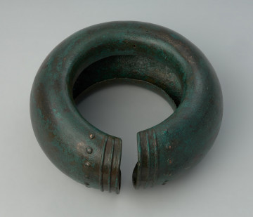 Pusty nagolennik z grubej brązowej blachy zdobiony na zakończeniach - Ujęcie z góry tyłu nagolennika. Pusty nagolennik z grubej brązowej blachy zdobiony na zakończeniach w postaci trzech poprzecznych żeberek oraz rzędu punktów. Od strony wewnętrznej widoczna szczelina.