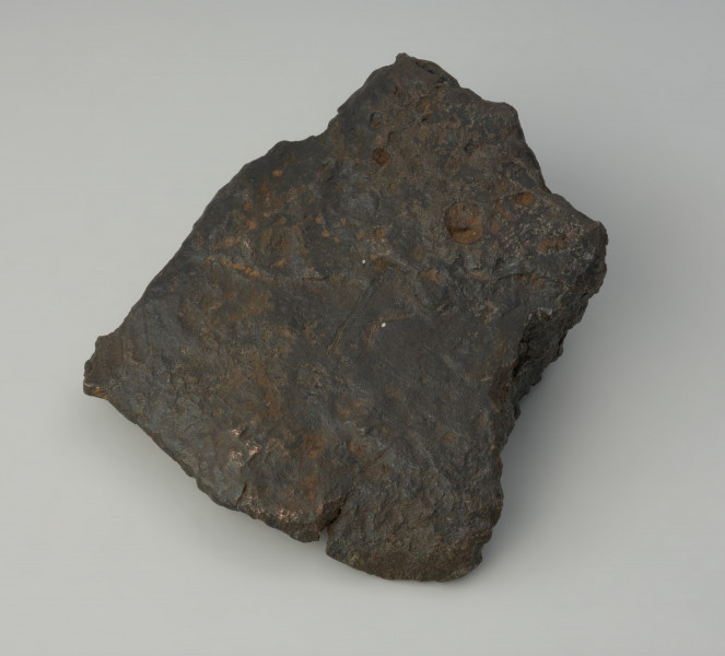 bryła metalu - Ujęcie w poziomie skosem w lewą stronę. Fragment placka odlewniczego o nieregularnym, czworobocznym kształcie z zachowanym jednym z pierwotnych boków o ostrej krawędzi. W przekroju poprzecznym trójkatny.