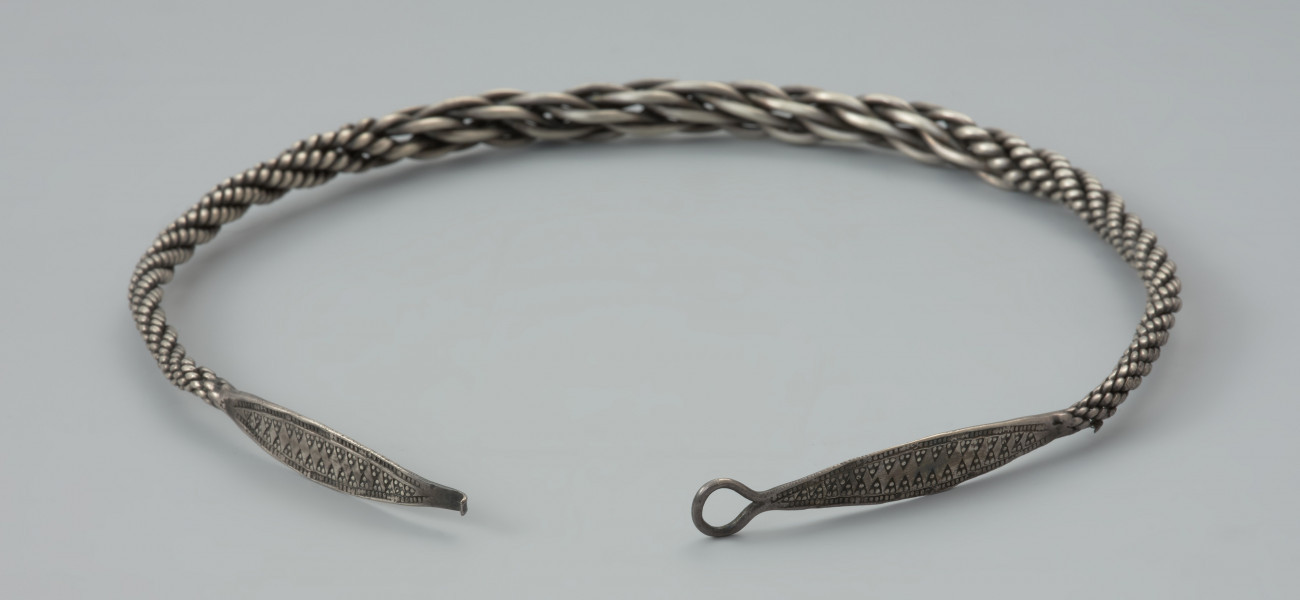 naszyjnik srebrny - Ujęcie zapięcia naszyjnika. Naszyjnik srebrny z czterech par skręconych drutów owijanych wokół siebie o grubości całkowitej od 0,4 do 1 cm. W części środkowej druty są skręcone zdecydowanie luźniej, co powoduje, że obręcz jest w tym miejscu grubsza. Oba końce obręczy są rozklepane w wydłużone owalne płytki zakończone uszkiem i haczykiem (fragment odłamany). Płytki są zdobione bordiurą z płaskich granulek wzdłuż obu krawędzi oraz dwoma rzędami trójkątów z trzema płaskimi granulkami w każdym z nich.