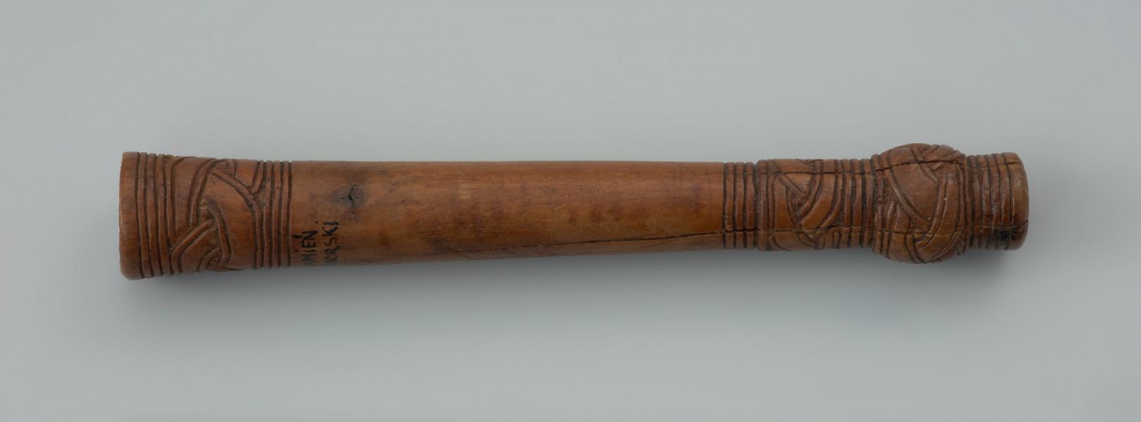 oprawka drewniana - Ujęcie z góry lewej strony oprawki. Drewniany trzonek podłużnego kształtu zbliżony do formy cylindrycznej. Na jednym z końców znajduje się kwadratowy otwór. Na obu przeciwległych stronach występują zdobienia w postaci prostej plecionki (warkocza) oraz dookolnych linii rytych. Zarówno trzonek jak i sam ornament wykonano z wielką starannością używając do tego ostrego noża.