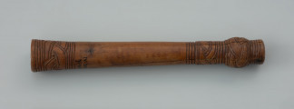 oprawka drewniana - Ujęcie z góry lewej strony oprawki. Drewniany trzonek podłużnego kształtu zbliżony do formy cylindrycznej. Na jednym z końców znajduje się kwadratowy otwór. Na obu przeciwległych stronach występują zdobienia w postaci prostej plecionki (warkocza) oraz dookolnych linii rytych. Zarówno trzonek jak i sam ornament wykonano z wielką starannością używając do tego ostrego noża.