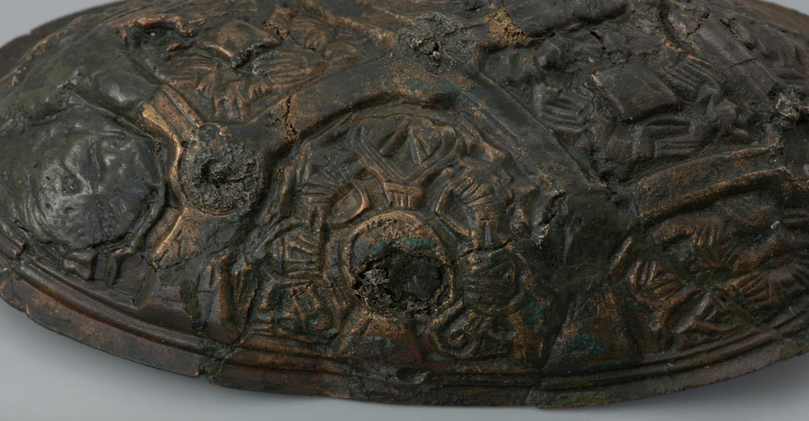 zapinka miseczkowata - Ujęcie fragmentu środkowej prawej części zapinki z góry. Zapinka ma kształt wydłużonego owalu. Przedmiot jest częściowo uszkodzony bez zachowanej szpili. Powierzchnia fibuli jest bogato ornamentowana motywami zoomorficznymi. Na detal zdobniczy składa się osiem pól wyznaczonych przez krzyżujące się wstęgi. W punktach ich przecięcia znajdują się ślady po siedmiu wypukłych guzkach. Na sześciu z ośmiu pól znajdują się motywy zwierzęce bliskie skandynawskiej sztuce w stylu Borre. Na trzech z czterech zachowanych pól zlokalizowanych w narożnikach fibuli występują zwierzęta chwytające swoimi szponami ciała innych stworzeń. Dwa kolejne naprzeciwległe pola, usytuowane w środkowej części odpowiadają dwóm łukowato wygiętym w biodrach zwierzętom - być może są to konie z głowami skierowanymi w swoją stronę. Pozostałe dwa pola zawierają przedstawienia zwierzęcych masek z połączonymi ze sobą zaciśniętymi łapami.