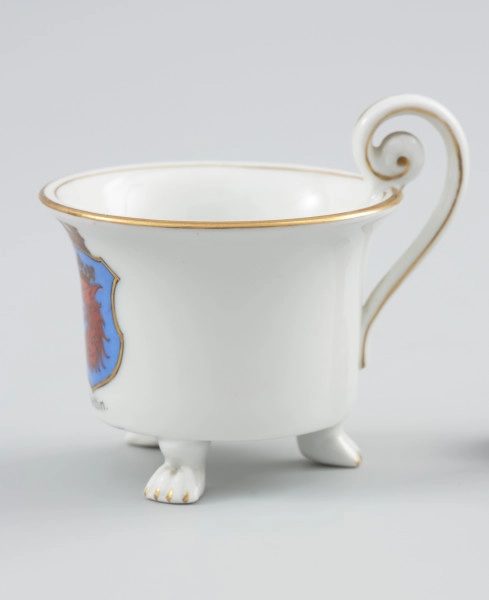 filiżanka w kształcie cylindrycznym - Ujęcie prawej strony. Filiżanka z białej porcelany o kształcie cylindrycznym, rozszerzającym się ku górze, na trzech nóżkach w formie łap zwierzęcych. Uszko wolutowe wysunięte ponad czarkę w obrębie zawinięcia woluty przymocowanej dołem do krawędzi czarki. Kształt uszka podkreślony w środku i na bocznych krawędziach złotym paskiem. Na ściance podszkliwna dekoracja - barwny herb Szczecina - niebieska tarcza ze złotym obrzeżem, o ściętych narożnikach, zwieńczona sepiowo-brązową koroną muralis. Wewnątrz tarczy czerwona głowa gryfa z czarnym rysunkiem piór, ze złotą koroną. Krawędź czarki opracowana złotą lamówką, powtórzoną wąskim paskiem wokół wewnętrznej krawędzi czarki. Nóżki, na których opiera się czarka, wyprofilowane w poczwórne pazury podkreślone złotym akcentem.