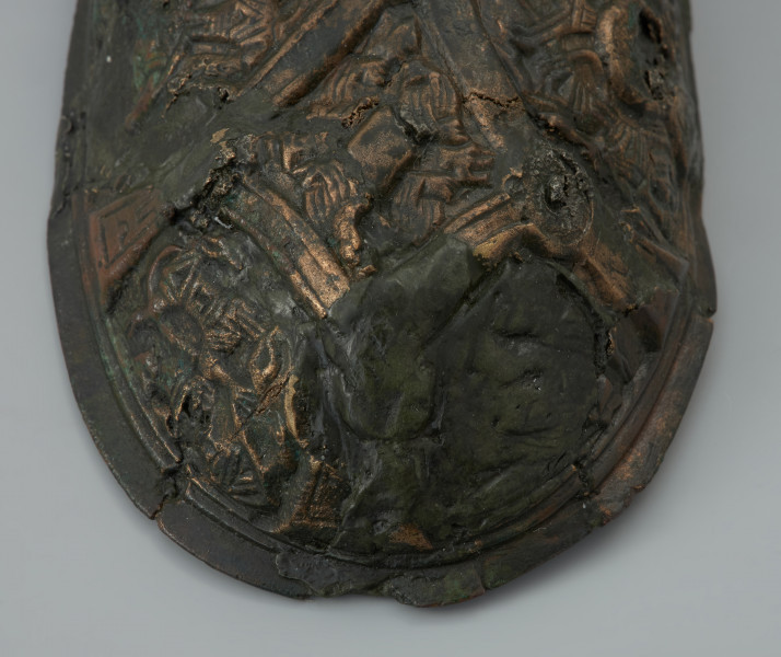 zapinka miseczkowata - Ujęcie fragmentu tylnej części zapinki z góry. Zapinka ma kształt wydłużonego owalu. Przedmiot jest częściowo uszkodzony bez zachowanej szpili. Powierzchnia fibuli jest bogato ornamentowana motywami zoomorficznymi. Na detal zdobniczy składa się osiem pól wyznaczonych przez krzyżujące się wstęgi. W punktach ich przecięcia znajdują się ślady po siedmiu wypukłych guzkach. Na sześciu z ośmiu pól znajdują się motywy zwierzęce bliskie skandynawskiej sztuce w stylu Borre. Na trzech z czterech zachowanych pól zlokalizowanych w narożnikach fibuli występują zwierzęta chwytające swoimi szponami ciała innych stworzeń. Dwa kolejne naprzeciwległe pola, usytuowane w środkowej części odpowiadają dwóm łukowato wygiętym w biodrach zwierzętom - być może są to konie z głowami skierowanymi w swoją stronę. Pozostałe dwa pola zawierają przedstawienia zwierzęcych masek z połączonymi ze sobą zaciśniętymi łapami.