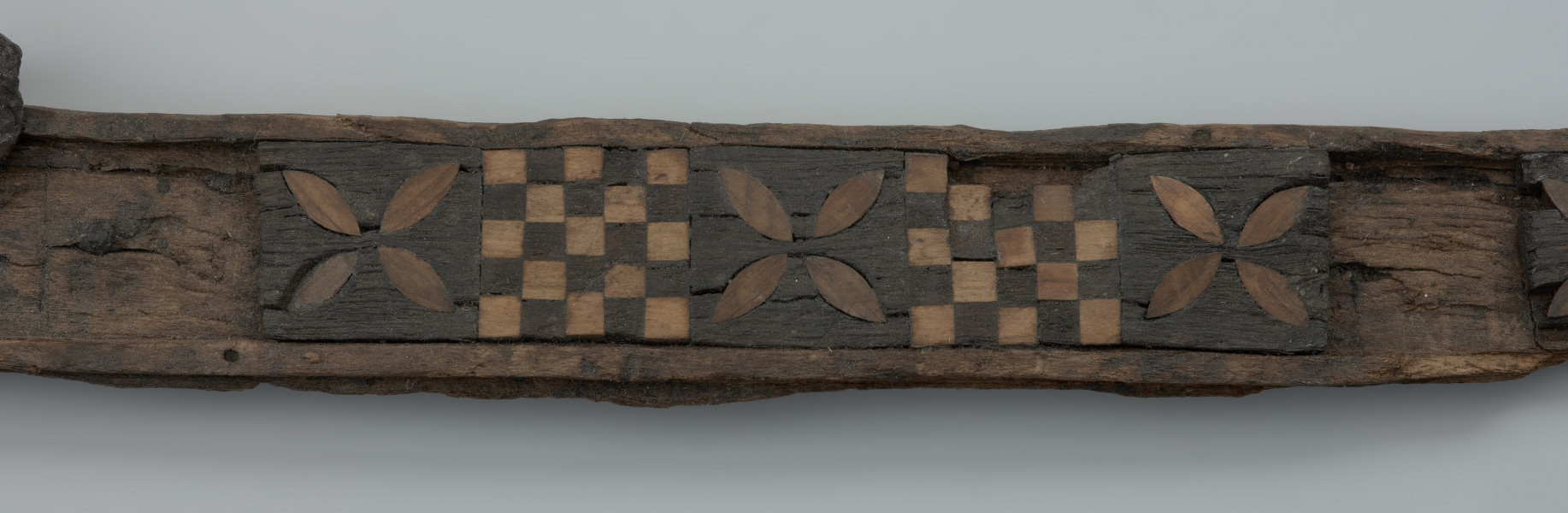 szkatułka intarsjowana - Ujęcie drugiego fragmentu szkatułki. Fragment wieko szkatułki z drewna bukowego, intarsjowana drewnem dębowym i cisowym.  Przybrzeżną część ścianki otacza kompozycja złożona z naprzemiennie umieszczanych motywów: szachownic o 25 polach i czterolistnych rozet z jasnego drewna, precyzyjnie wkomponowanych w soczewkowate wycięcia w płycinach z ciemniejszej dębiny.