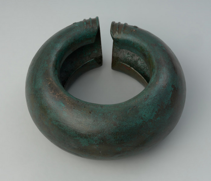 Pusty nagolennik z grubej brązowej blachy zdobiony na zakończeniach - Ujęcie z tgóry przodu nagolennika. Pusty nagolennik z grubej brązowej blachy zdobiony na zakończeniach w postaci trzech poprzecznych żeberek oraz rzędu punktów. Od strony wewnętrznej widoczna szczelina.