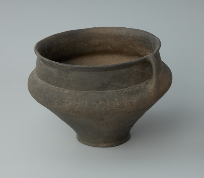 Ujęcie z boku. Waza gliniana z uchem o podciętej dolnej partii brzuśca i wysokim załomie. Szyja naczynia jest niemal cylindryczna, wylew delikatnie wychylony na zewnątrz i zaokrąglony. Waza jest ciebnobrunatna, o wygładzanej powierzchni. Naczynie jest zdobiona dookolnym pasem ornamentacyjnym w postaci powielonych swastyk wykonanym kółkiem zębatym.