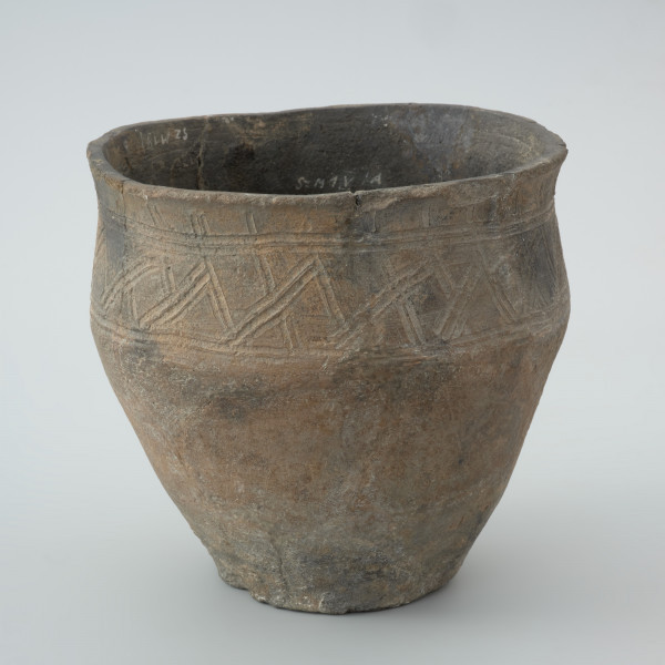 naczynie gliniane - Ujęcie z prawej strony. Garnek ma kształt dwustożkowaty o esowatym profilu z charakterystyczną wychyloną na zewnątrz krawędzią wylewu oraz łagodny załom brzuśca znajdujący się w 2/3 wysokości. Na powierzchni pojemnika powyżej załomu występuje zdobienie w postaci krzyżujących się skośnych linii wpasowanych w podwójne, dookolne linie ryte. Naczynie wykonano w technice częściowego obtaczania na kole garncarskim. Uformowano je poprzez sukcesywne klejenie glinianych wałeczków albo taśm. W centralnej części dna znajduje się wklęsły odcisk po ośce, na której obracało się koło. Do produkcji wykorzystano masę garncarską z domieszką schudzającą drobno i średnioziarnistego tłucznia w postaci piasku i granitu.