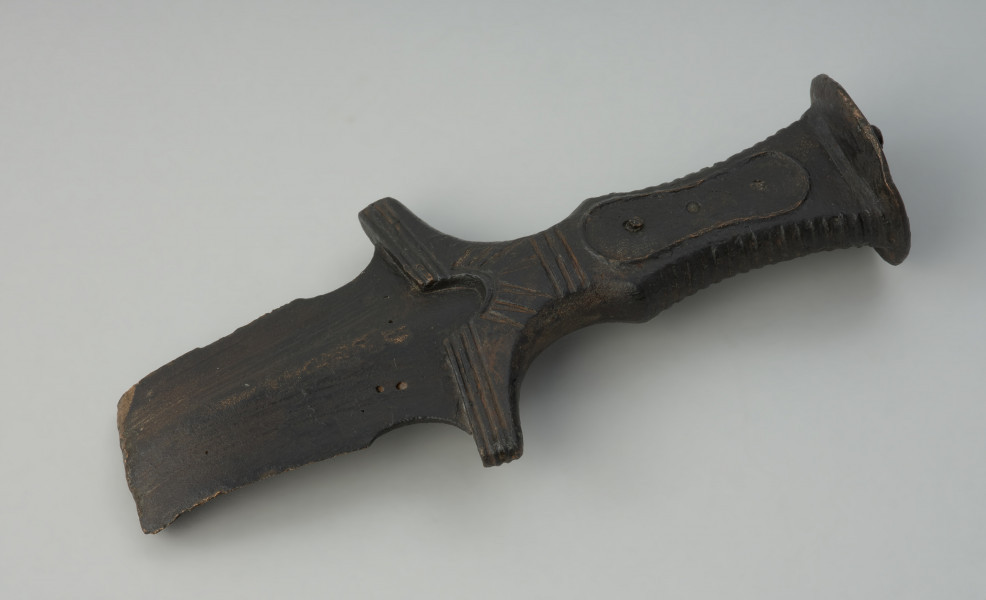 broń, miecz - Ujęcie w poziomie z przodu skosem w lewą stronę. Brązowy miecz z czworokatną, dekorowaną żłobieniami rękojeścią, głowicą w formie owalnej tarczki oraz jelcem ze skrzydełkami i półkolistym wycięciem.