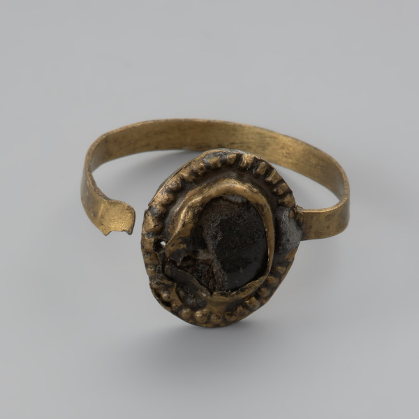 pierścionek z oczkiem - Ujęcie z przodu. Trzyczęściowy brązowy pierścionek z oczkiem, składający się z metalowej taśmy stanowiącej otok, metalowej tarczki pod oczko złożonej z owalnej płaskiej blaszki spodniej oraz dodatkowej taśmy tworzącej eliptyczne gniazdko. Jest ona zdobiona dookoła odciskami. Trzecim elementem, który uzupełnia oczko jest grafitowe szkiełko zachowane jedynie w części, przyklejone do spodniej tarczki i przytrzymywane przez krawędzie otaczającej je taśmy. Między blaszkami oczka pozostawione są dwie 