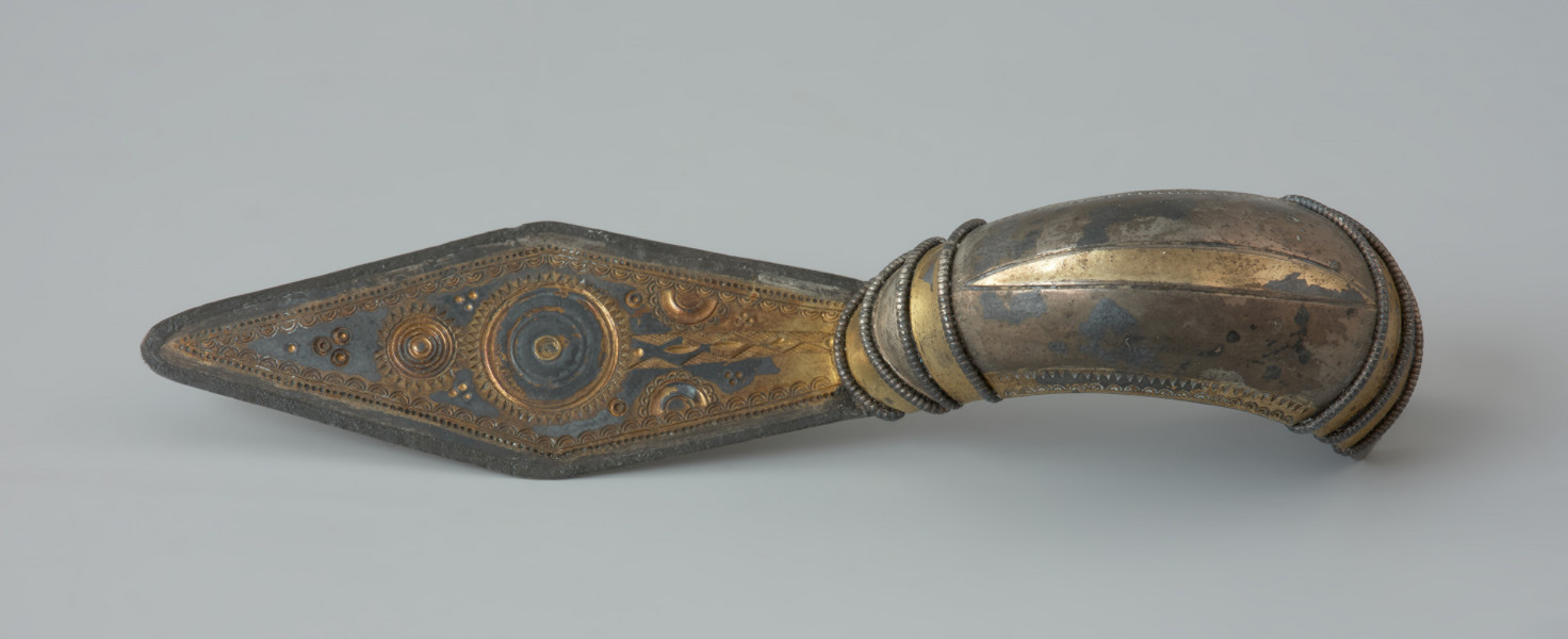 Ujęcie z góry. Srebrna zapinka (kabłąk) pozłacana o długiej rombowatej nóżce. Kabłąk jest zdobiony na końcach poprzecznie grupami 3 i 4 odcinków perełkowatego drucika połączonymi poziomo analogicznymi łukowatymi fragmentami takich drucików.