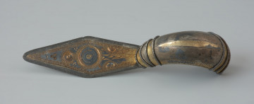 Ujęcie z góry. Srebrna zapinka (kabłąk) pozłacana o długiej rombowatej nóżce. Kabłąk jest zdobiony na końcach poprzecznie grupami 3 i 4 odcinków perełkowatego drucika połączonymi poziomo analogicznymi łukowatymi fragmentami takich drucików.