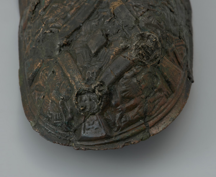 zapinka miseczkowata - Ujęcie fragmentu przedniej części zapinki z góry. Zapinka ma kształt wydłużonego owalu. Przedmiot jest częściowo uszkodzony bez zachowanej szpili. Powierzchnia fibuli jest bogato ornamentowana motywami zoomorficznymi. Na detal zdobniczy składa się osiem pól wyznaczonych przez krzyżujące się wstęgi. W punktach ich przecięcia znajdują się ślady po siedmiu wypukłych guzkach. Na sześciu z ośmiu pól znajdują się motywy zwierzęce bliskie skandynawskiej sztuce w stylu Borre. Na trzech z czterech zachowanych pól zlokalizowanych w narożnikach fibuli występują zwierzęta chwytające swoimi szponami ciała innych stworzeń. Dwa kolejne naprzeciwległe pola, usytuowane w środkowej części odpowiadają dwóm łukowato wygiętym w biodrach zwierzętom - być może są to konie z głowami skierowanymi w swoją stronę. Pozostałe dwa pola zawierają przedstawienia zwierzęcych masek z połączonymi ze sobą zaciśniętymi łapami.