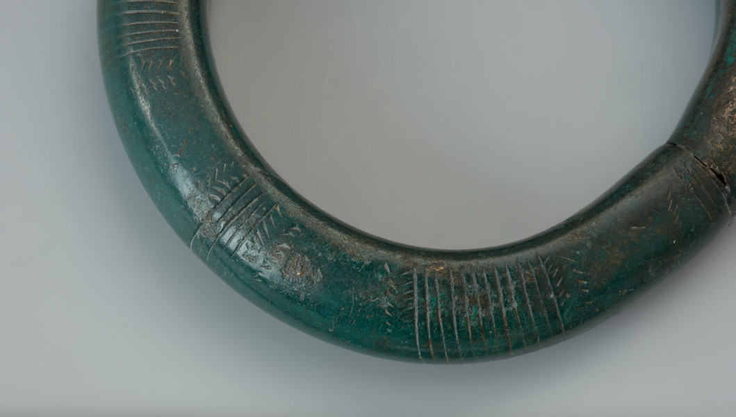 Bransoleta obręczowa - Powiększenie przodu bransolety ze zdobieniami. Masywna bransoleta wykonana z grubego brązowego pręta o przekroju kolistym. Końce obręczy są ścięte prosto i ściśle do siebie przylegaja. Powierzchnię zabytku pokrywa delikatnie wyryte zdobienie złożone z grup poprzecznych kresek, połączonych z motywem jodełki. Bransoletę pokrywa ciemnozielona patyna.