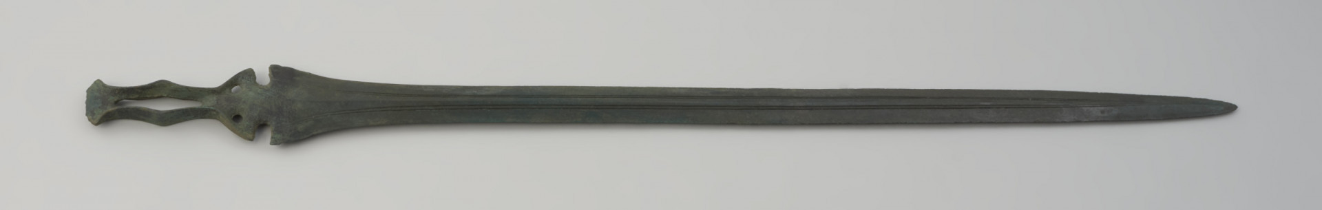 miecz ze sztabą do rękojeści - Ujęcie całego miecza w poziomie z przodu. Zachowany w całości miecz obosieczny o smukłej głowni z widocznymi podwójnymi struzinami na powierzchni. Sztaba do rękojeści ma oryginalną formę, poszerzoną pośrodku i zwieńczoną wachlarzowatym zakończeniem. Dodatkowo na całej jej długości przebiega charakterystyczna, rombowata szczelina. Pókolista podstawa sztaby zaopatrzona jest w 2 otwory na nity. Obydwie powierzchnie miecza są lekko porowate i pokryte zieloną patyną.