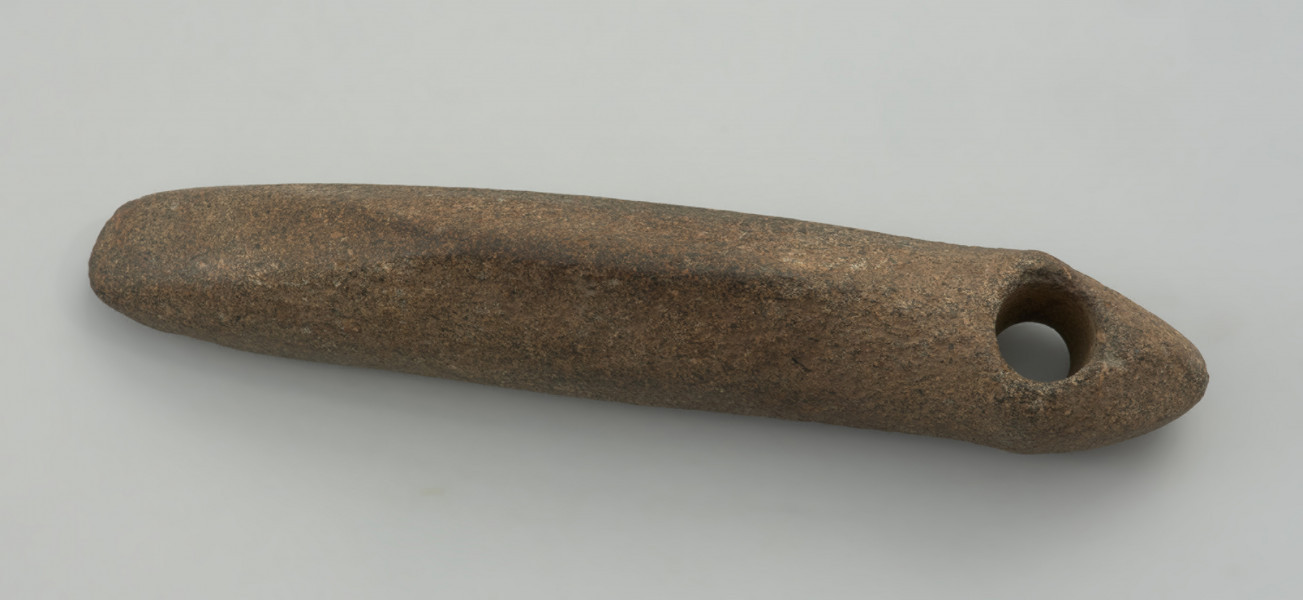 narzędzie, motyka - Ujęcie z przodu;  Smukłe narzędzie o ostrzu poprzecznym (motyka), o bokach prostych w rzucie z góry, z wyodrębnioną częścią tylną z otworem i spiczastym zakończeniem w formie przypominającej główkę żmii. Otwór jest wywiercony jednostronnie. Powierzchnia gładzona, ma fakturę od gładkiej do lekko szorstkiej.
