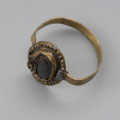 pierścionek z oczkiem - Ujęcie z góry przodu skosem w lewą stronę. Trzyczęściowy brązowy pierścionek z oczkiem, składający się z metalowej taśmy stanowiącej otok, metalowej tarczki pod oczko złożonej z owalnej płaskiej blaszki spodniej oraz dodatkowej taśmy tworzącej eliptyczne gniazdko. Jest ona zdobiona dookoła odciskami. Trzecim elementem, który uzupełnia oczko jest grafitowe szkiełko zachowane jedynie w części, przyklejone do spodniej tarczki i przytrzymywane przez krawędzie otaczającej je taśmy. Między blaszkami oczka pozostawione są dwie 