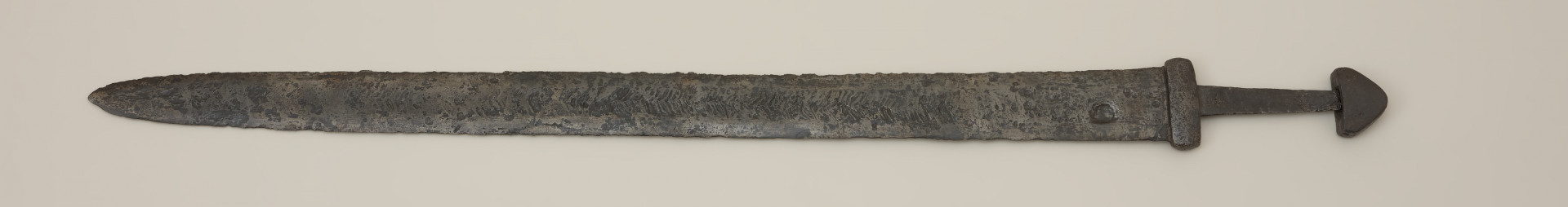 broń; miecz dwusieczny - Ujęcie przodu miecza. Całkowicie zachowany. Dwusieczna głownia jest szeroka i masywna, o mniej więcej jednakowej szerokości na całej długości, przy zastawie ma przekrój sześcioboczny. Sztych jest zaokrąglony. Na obu płazach głowni widoczny wzór w postaci „jodełki” świadczący o wieloelementowej jej konstrukcji. Rdzeń został wykonany z dwóch skręconych prętów, nisko- i wysokowęglowego, tworzących jądro, z dodanymi po obu stronach nakładkami ostrzy. Trzpień miecza jest wąski i lekko zwęża się ku głowicy. Jelec jest krótki i wysoki, o kształcie widzianym od góry zbliżonym do prostokąta o lekko wypukłych krótszych bokach. Głowica jest jednolita, zbliżona do trójkąta. Jelec i głowica są lite, wykute każde z pojedynczego kawałka metalu.