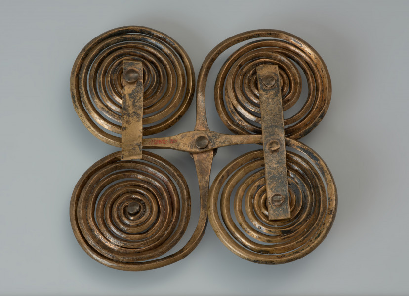 Zapinka krzyżowa - Ujęcie z dołu. Zapinka krzyżowa wykonana jest z dwóch brązowych prętów o rombowatym przekroju, z których uformowano po parze spiralnych tarczek, nadając im ósemkowate kształty. Czworokątny kształt uzyskano składajac na krzyż obydwa elementy i spajając je od spodu dwoma wąskimi blaszkami, z których jedna zachowana jest w połowie. Fibulę udekorowano pięcioma ozdobnymi guzkami.