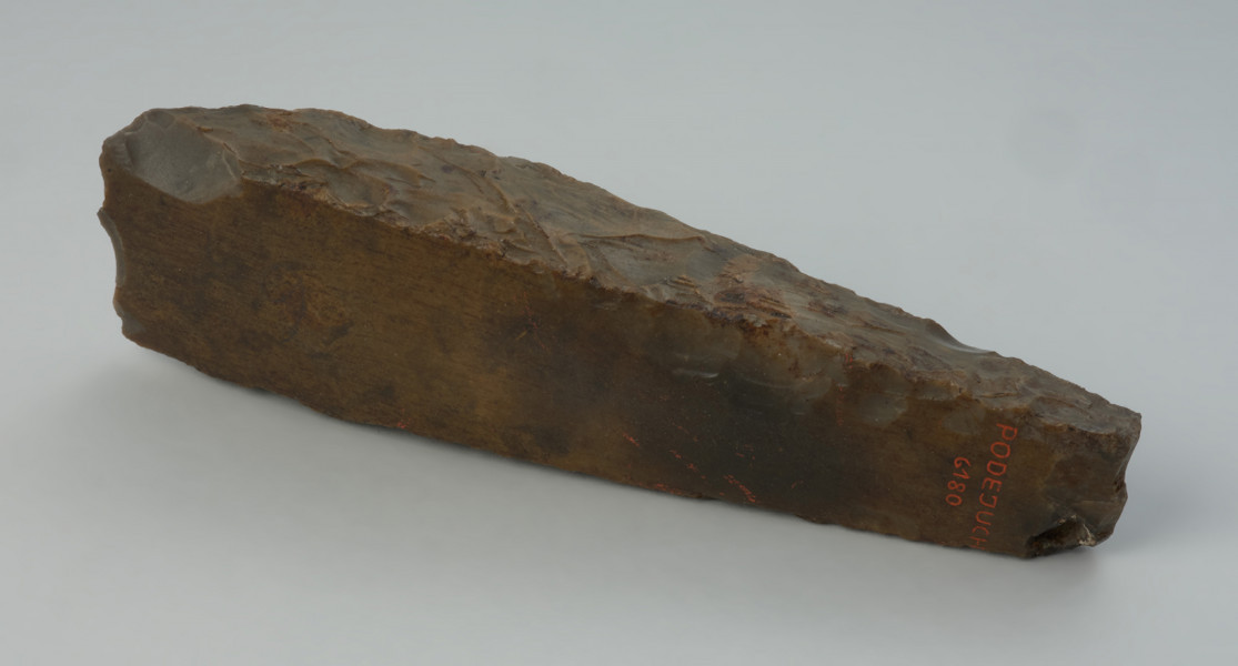 narzędzie - Ujęcie z boku; Smukła, długa ciosła czworościenna o trapezowatym kształcie, z wąskim, tępym obuchem. W przekroju poprzecznym prostokątne, przy czym bliżej ostrza dłuższe boki są łukowate: górny wklęsły, a dolny wypukły, co pozwalało ukształtować łukowatą krawędź ostrza. Samo ostrze jest zniszczone. Powierzchnia szlifowana z widocznymi śladami odnawiania narzędzia w postaci negatywów (retuszu) na powierzchni górnej, w części od połowy długości do obucha na ściance górnej. Przy obuchu, który jest ukształtowany nieregularnie, znajduje się naturalne zagłębienie pokryte korą wapienną. Cała powierzchnia szlifowana pokryta jest jasnobrunatną patyną. Części negatywowe są spatynowane znacznie słabiej. Przedmiot został wykonany z krzemienia kredowego o szarej barwie.