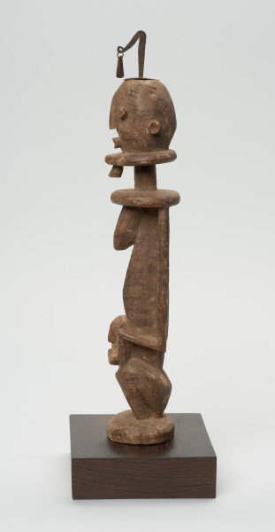 rzeźba - Ujęcie lewego boku. Drewniana, rzeźbiona Postać z dzieckiem na kolanach. Siedzi w pozycji przykucniętej. Głowa i tułów nieproporcjonalnie duże w stosunku do nóg. Oczy, nos, usta, uszy mocno wypukłe. Dziecko ułożone na kolanach, zasłania twarz rękoma. Na głowie fragment żelaza.