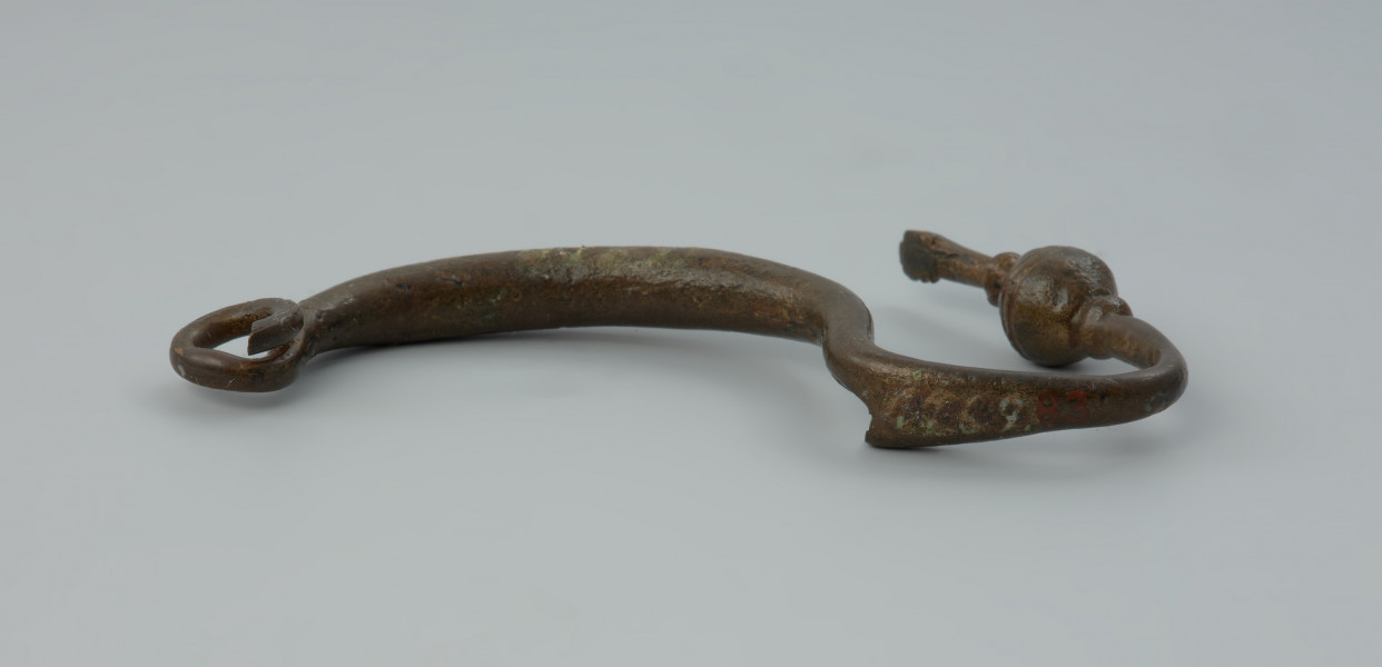 Kabłąk zapinki z brązu - Ujęcie z tyłu. Zapinka z brązu; zachował się tylko kabłąk jednoczęściowy, przechodzący w odgiętą w górę nóżkę, zachowany jest jeden zwój sprężynki.