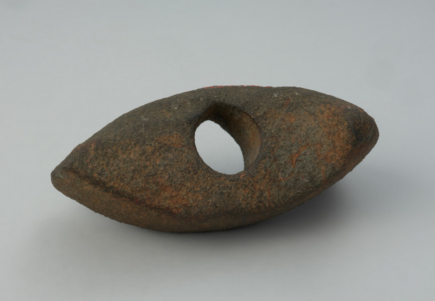 Topór kamienny - Ujęcie z góry; Topór z szerokim, symetrycznie rozszerzonym ostrzem i nieznacznie rozszerzonym obuchem, o prostokątnym przekroju poprzecznym, wykonany z szarego surowca skalnego. Otwór niemal kolisty, cylindryczny. Powierzchnia silnie zerodowana.