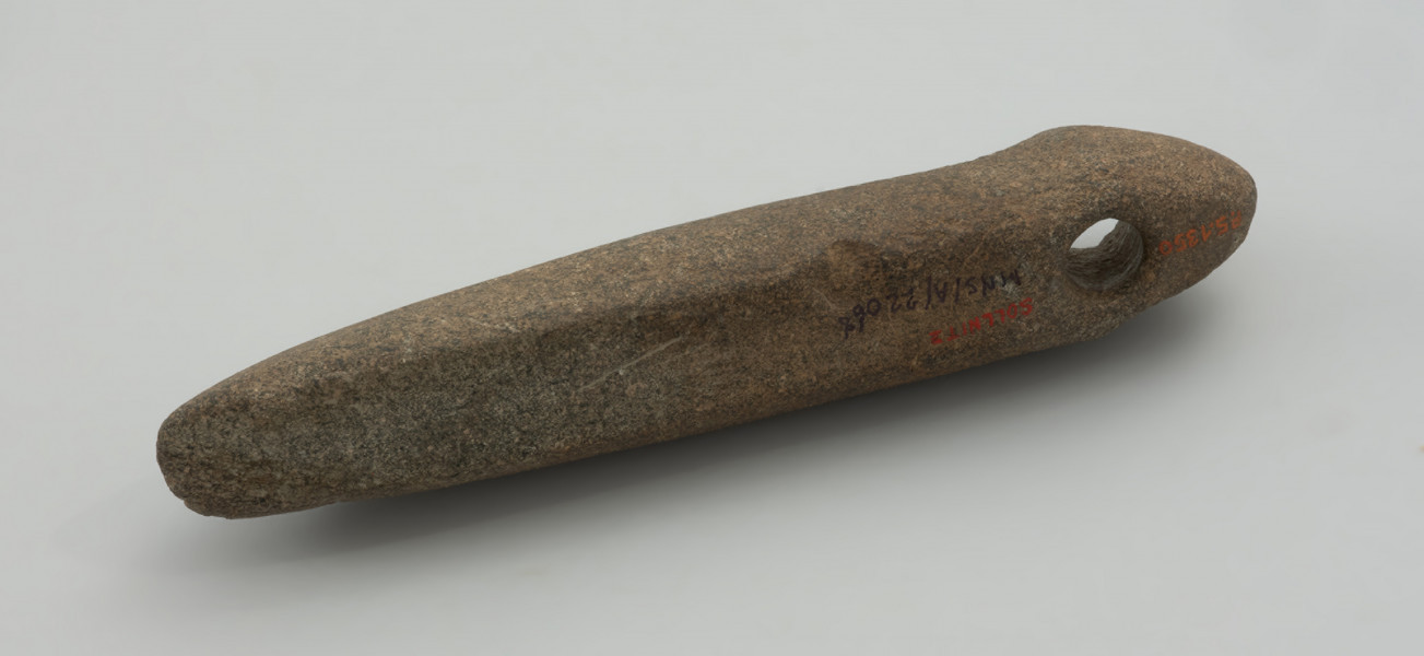 narzędzie, motyka - Ujęcie z tyłu; Smukłe narzędzie o ostrzu poprzecznym (motyka), o bokach prostych w rzucie z góry, z wyodrębnioną częścią tylną z otworem i spiczastym zakończeniem w formie przypominającej główkę żmii. Otwór jest wywiercony jednostronnie. Powierzchnia gładzona, ma fakturę od gładkiej do lekko szorstkiej.