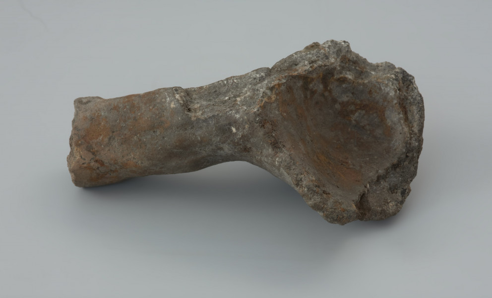 Łyżka odlewnicza - Ujęcie prawego boku. Łyżka odlewnicza wykonana z bardzo mocno spieczonej gliny zachowana fragmentarycznie w postaci części okrągłej, głebokiej nabierki i fragmentu masywnego trzonka