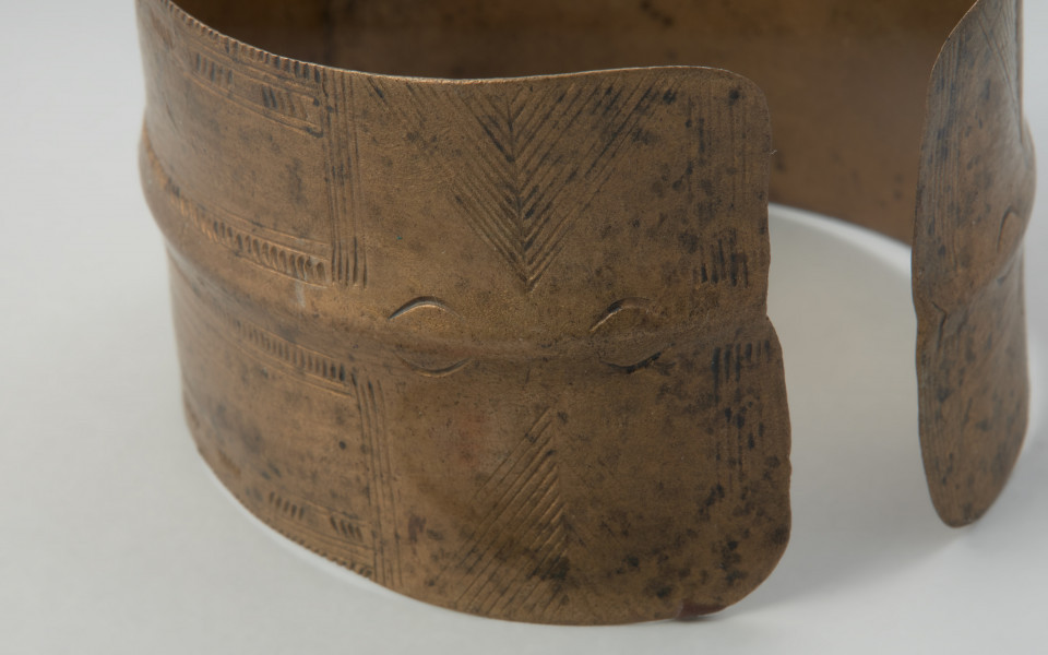 bransoleta taśmowata - Ujęcie prawego boku bransolety. Bransoleta otwarta wykonana z wąskiej, dość grubej taśmy brązowej o przekroju płasko-wypukłym. Końce obręczy są ścięte prosto, a jej powierzchnię pokrywa ornament ryty złożony z grup ukośnych i pionowych kresek, poprzedzielanych motywem jodełki.