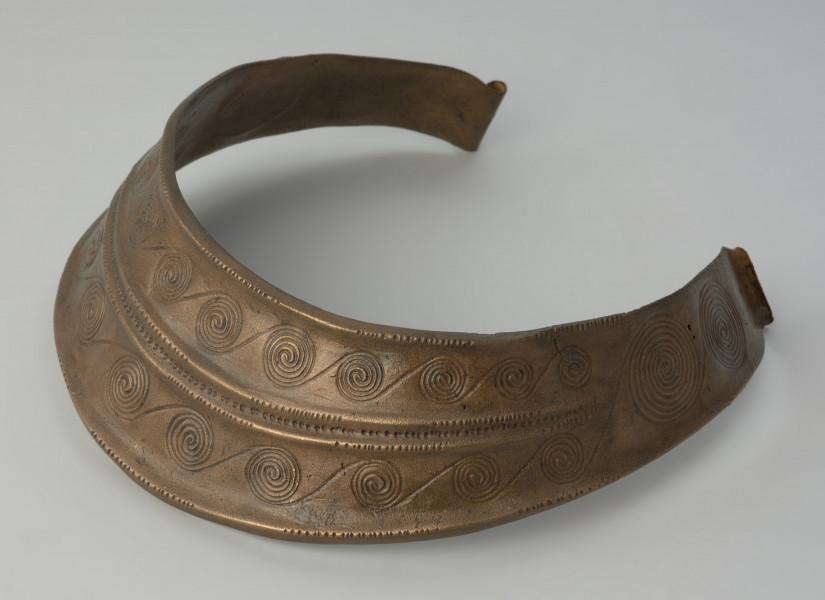 Brązowy naszyjnik kołnierzowaty - Ujęcie zprawego boku. Brązowy naszyjnik kołnierzowaty jest zdobiony motywem  spiralnych pasm ułożonych w dwóch rzędach, oddzielonych podwójnym żeberkiem, ornamentowanym
drobnymi nacięciami i punktowaniem.