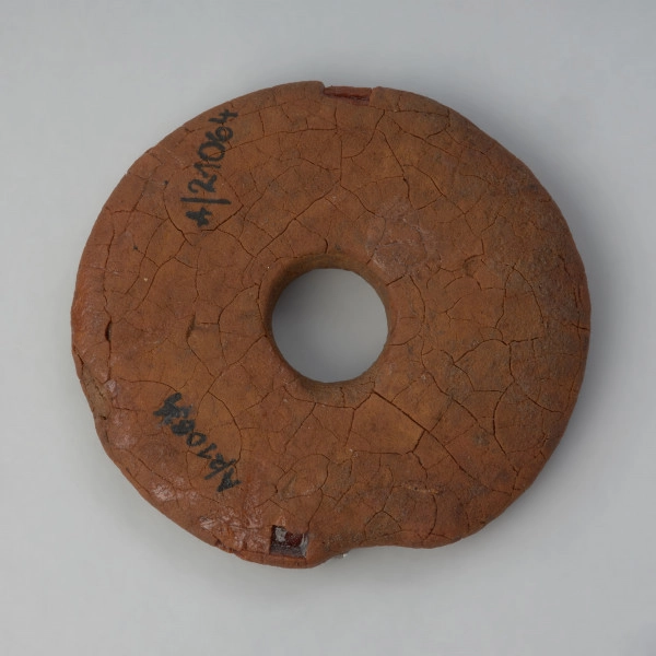 zawieszka, amulet - Ujęcie z góry spodu tarczki. Tarczka bursztynowa w formie dysku z niewielkim otworem pośrodku o spatynowanej i popękanej powierzchni.