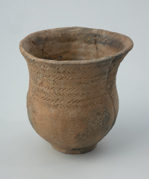Naczynie gliniane - Ujęcie z przodu; Przysadzisty puchar o szerokim wylewie, zdobiony na szyjce czterema dookolnymi szeregami ukośnych, podłużnych odcisków, które tworzą pasma złożone z dwóch szeregów odcisków układających się w motyw rybich ości. Ukośnymi nacięciami jest zdobiona także krawędź wylewu.